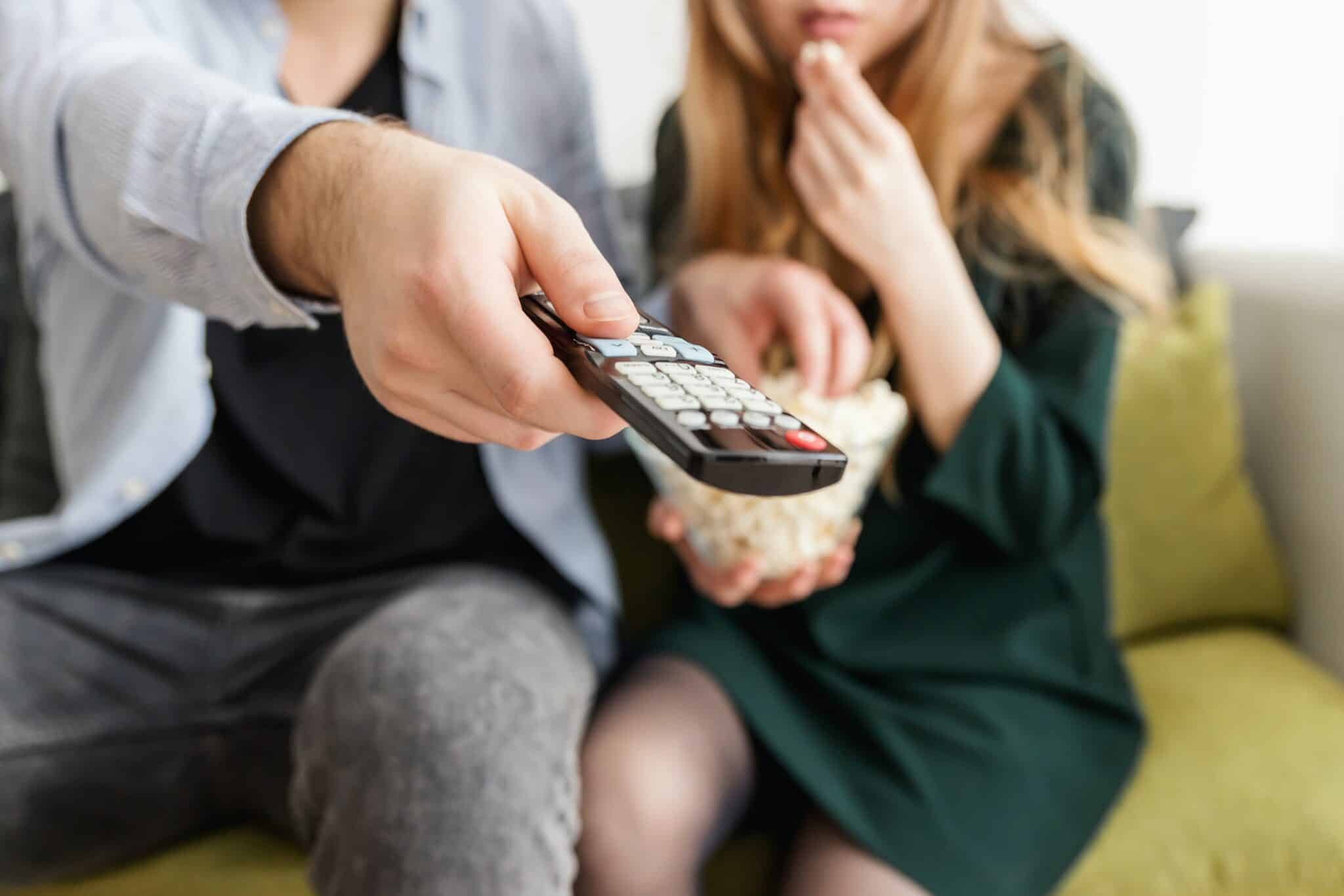 Couple watching TV at home with man pointing remote at the TV.
